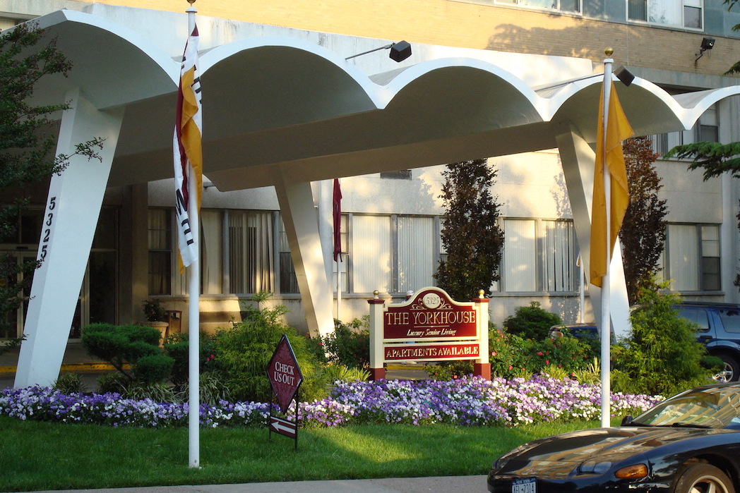 In front of the awning to the building's entrance are flags, a flowerbed and signage.